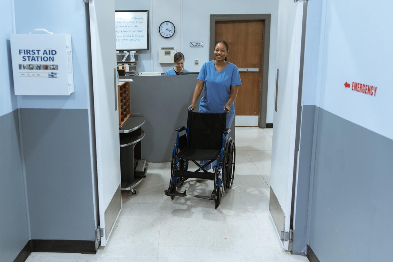 A woman in blue scrubs pushing a wheelchair down the hallway.