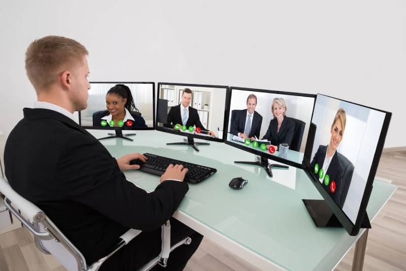 A man in black jacket sitting at table with four computers.